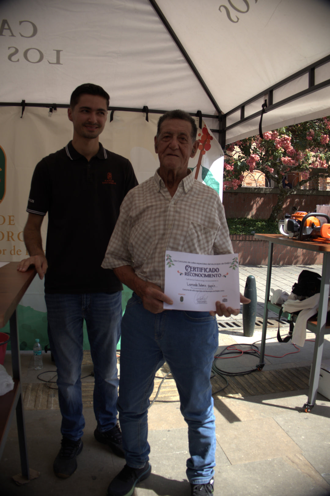 Two men holding a certificate outdoors with a tent and floral decorations in the background